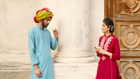 Hindu-happy-woman-with-dot-on-forehead-and-man-in-turban-standing-outdoors,-talking-and-drinking-coffe-or-tea.-Resting.-Handsome-male-and-beautiful-female-sipping-drink-and-spending-time-together.